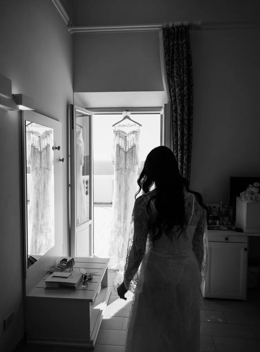 Bride in a robe approaching her wedding dress hanging in the balcony doorway, black and white