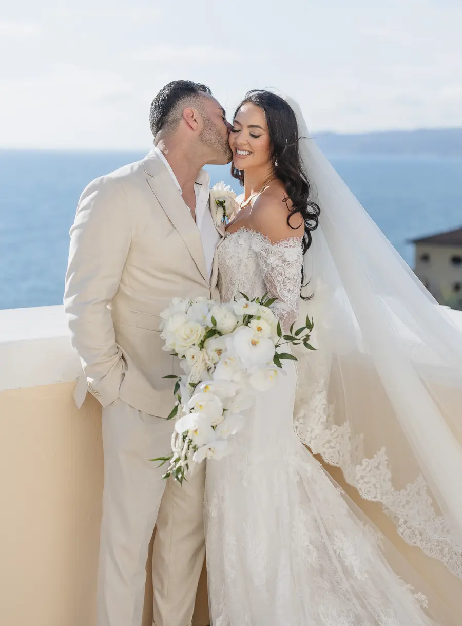 Groom kissing the bride's cheek, orchid bouquet and lace veil by the sea