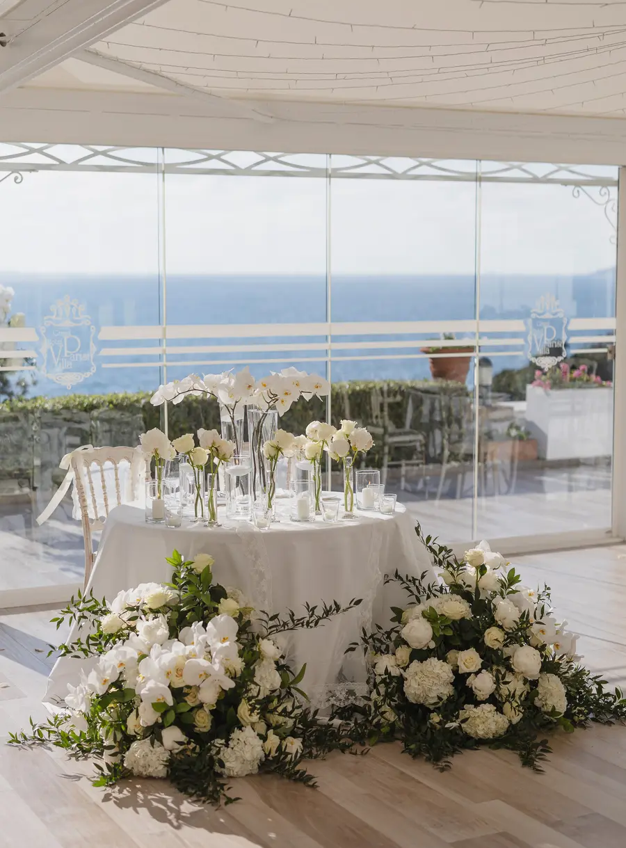Sweetheart table with orchid vases and white flowers in the glass pavilion by the sea