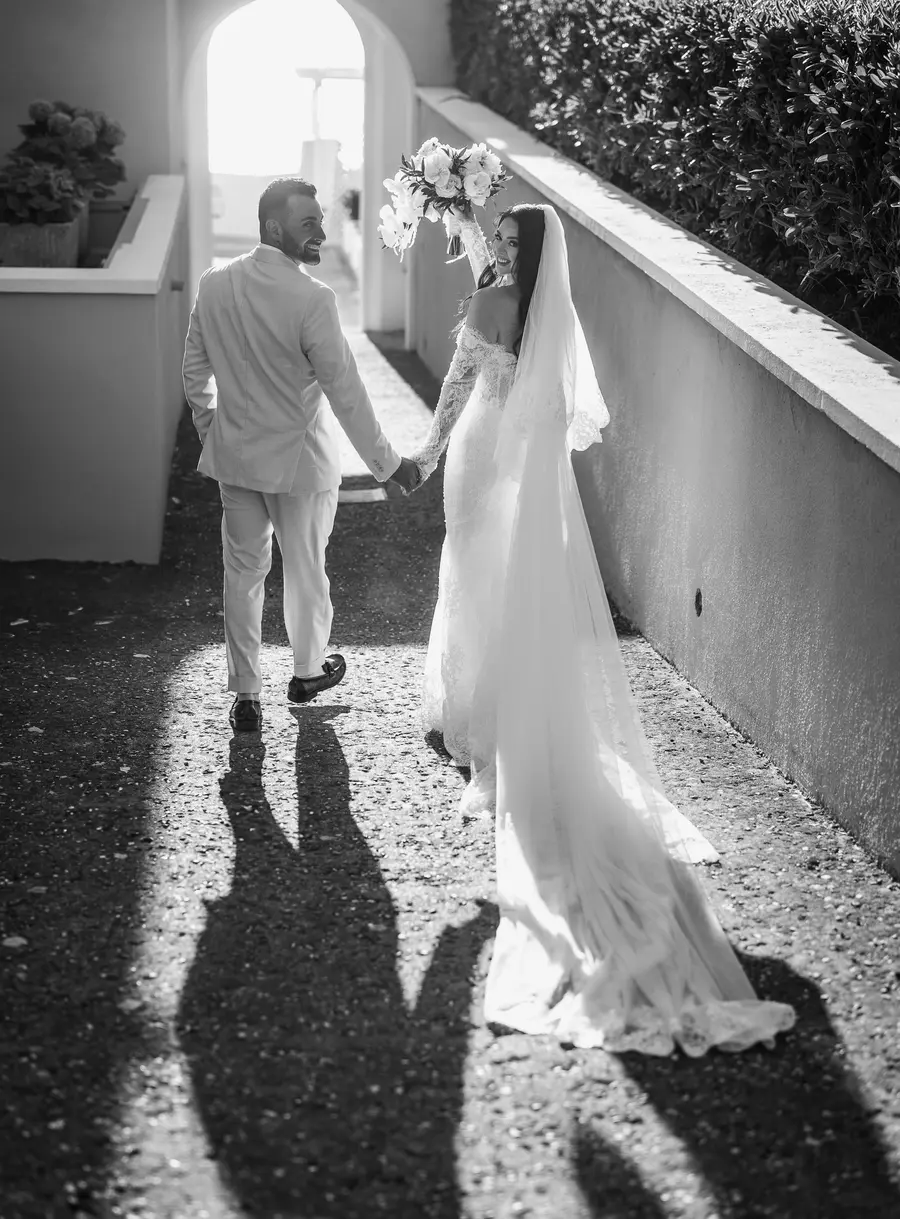 Newlyweds hand in hand along the backlit path, the bride looking back, black and white