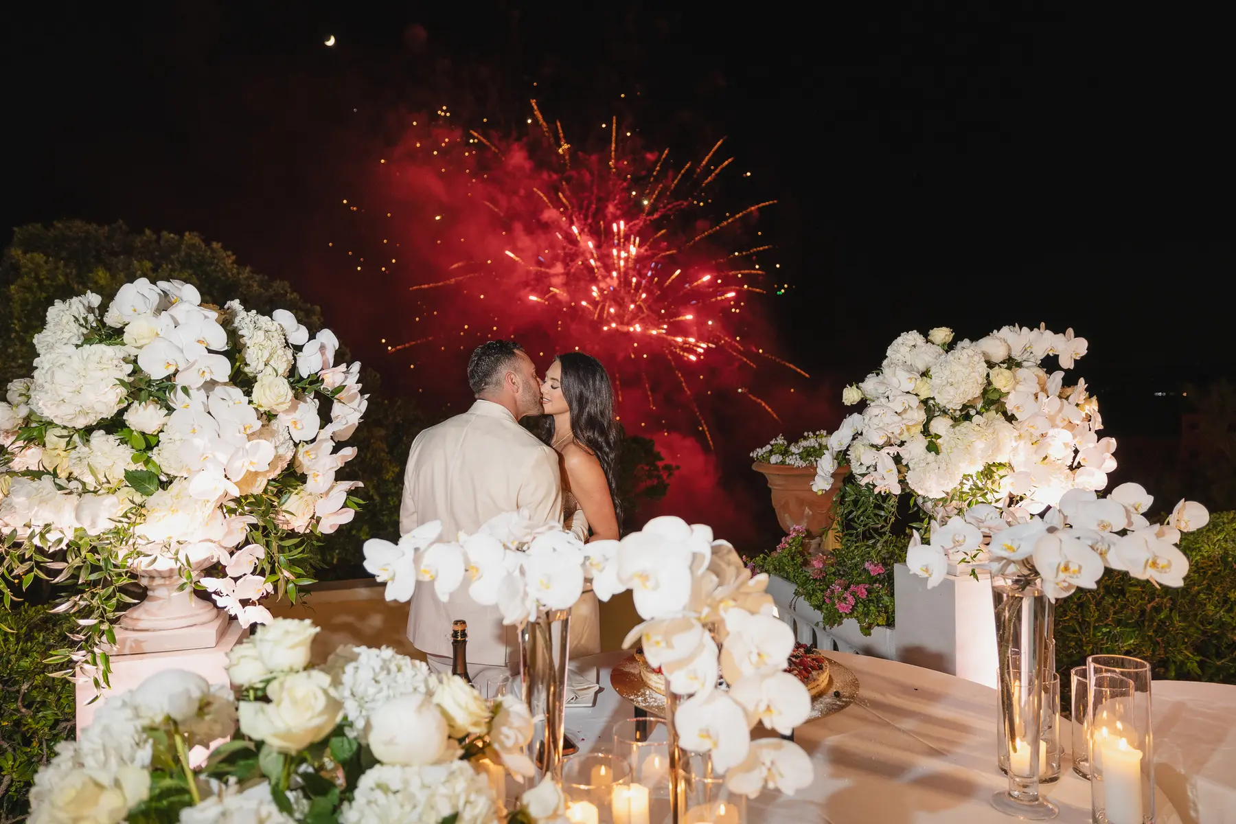 Couple kissing at night among white flowers as fireworks light up the sky