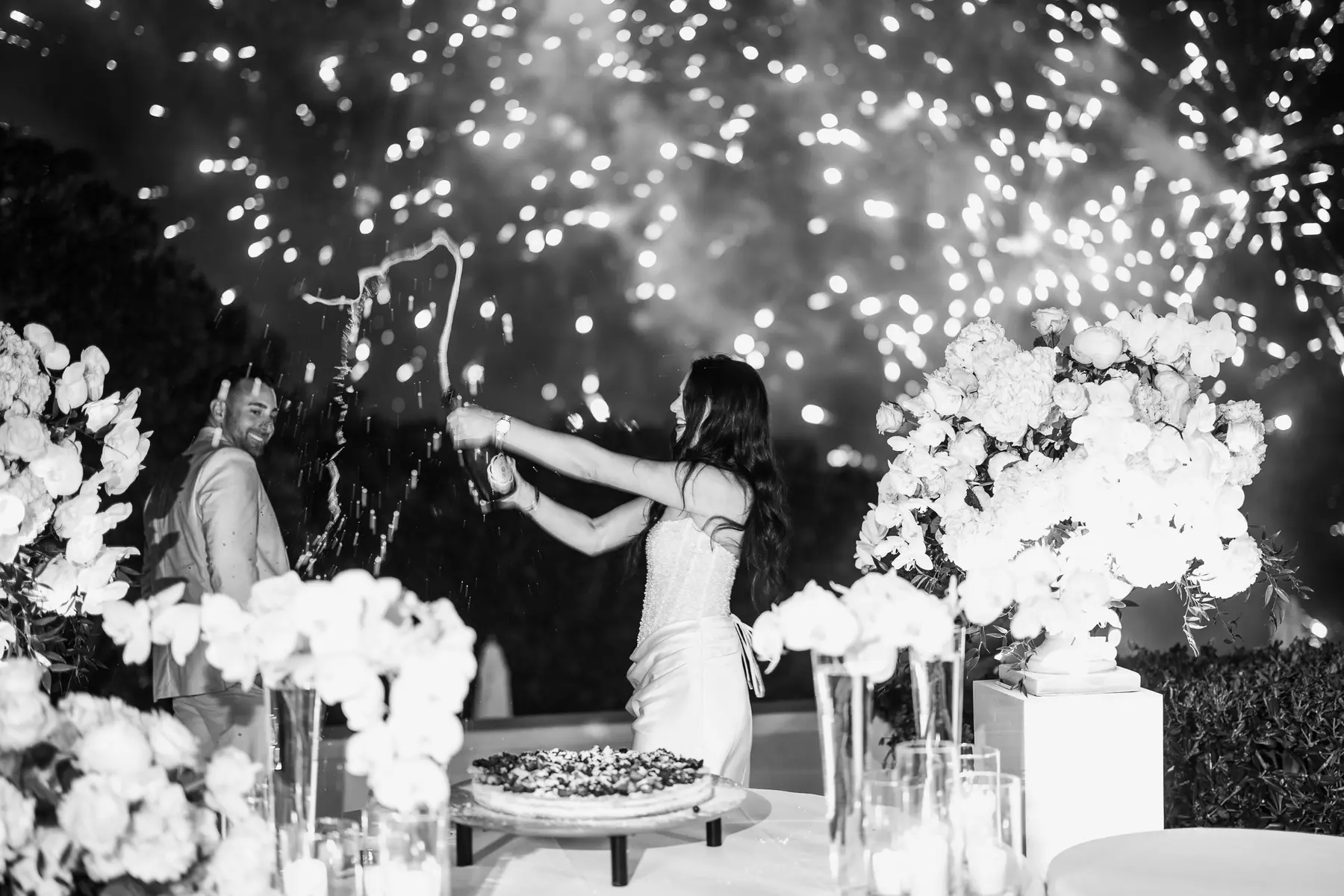 Bride spraying sparkling wine amid fireworks beside the wedding cake, black and white